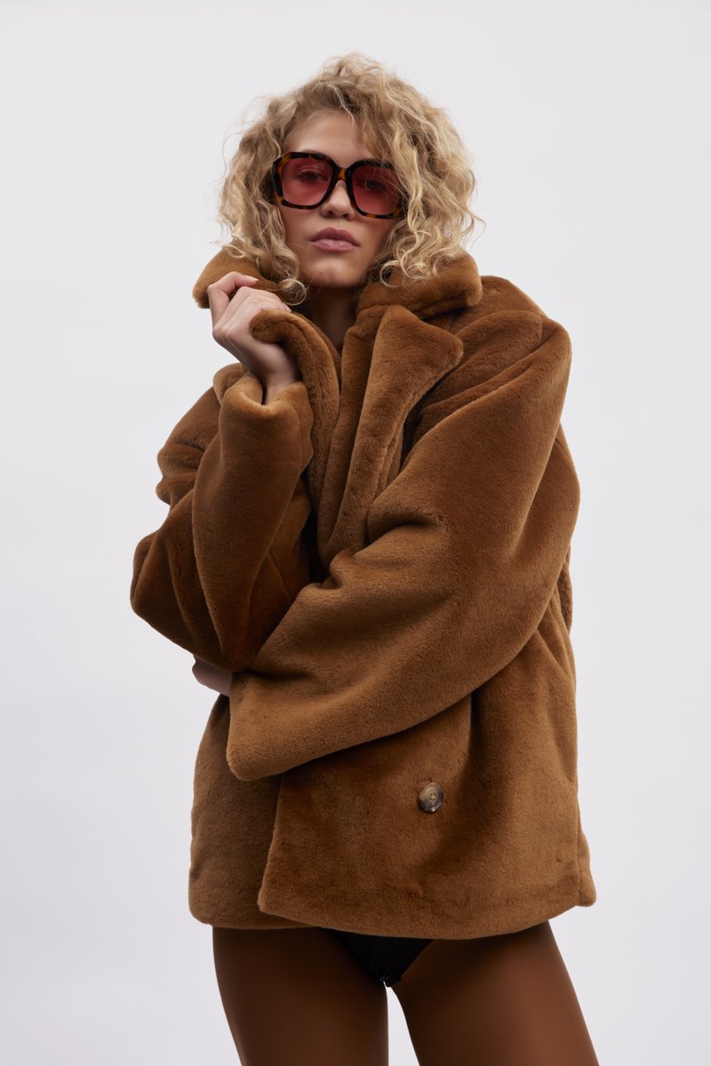 Brun kunstlær frakk med bred krage og oversized passform. Har enkelt knappefeste og myk tekstur. Et par oversized rosa solbriller.