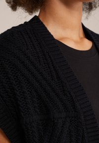 Cardigan negro de punto con una textura gruesa y un diseño de frente abierto. Presenta bordes acanalados y un patrón en zigzag a lo largo del tejido.