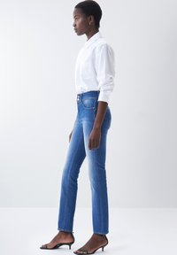 Los jeans de mezclilla azules ajustados presentan una cintura alta y un diseño desgastado. Se combinan con una camisa blanca de botones y sandalias negras de tacón.