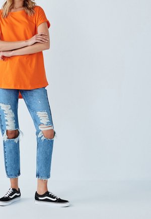 Personne portant un haut orange à manches courtes, un jean bleu déchiré et des baskets décontractées noires et blanches, debout les bras croisés.