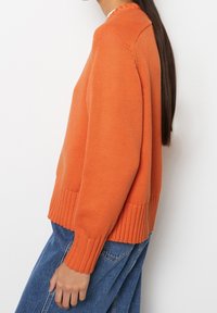 Maglione arancione lavorato a maglia con polsini e orlo a coste, caratterizzato da una vestibilità comoda e un tessuto texturizzato. Abbinato a denim blu.