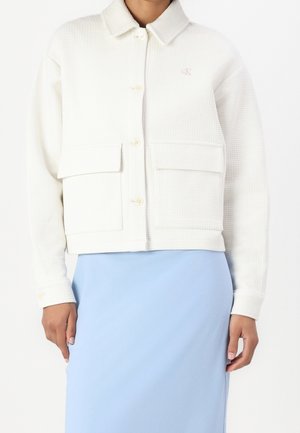 Woman wearing a white textured button-up jacket with two oversized front pockets and a light blue skirt.