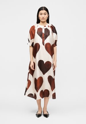 Žena stojaca v bielej midi šaty s veľkými hnedými srdcovými potlačami, nafúknutými rukávmi, gombíkovým výstrihom a čiernymi špicatými balerínami.