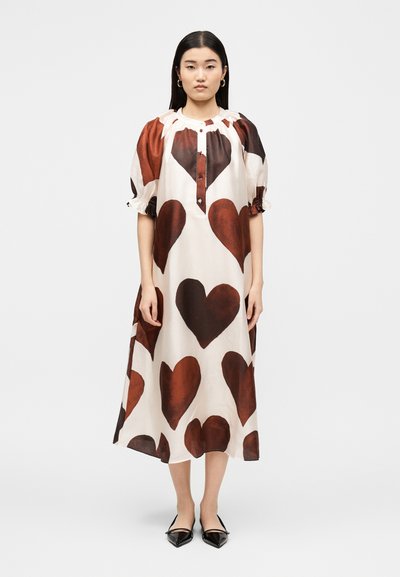 Woman standing wearing a white midi dress with large brown heart prints, puffed sleeves, buttoned neckline, and black pointed flats.
