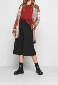Giacca a camicia oversize a quadri arancione e marrone, abbinata a un maglione rosso testurizzato, culotte nere e stivali neri alla caviglia.