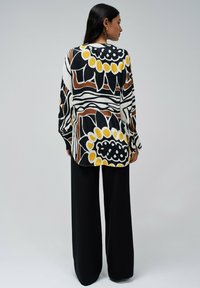 Chemise à manches longues avec un motif floral et abstrait multicolore en noir, blanc, jaune et marron, associée à un large pantalon noir.