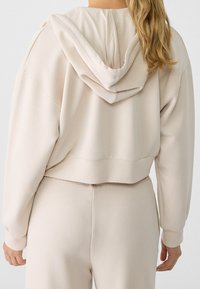 Sudadera corta de color beige claro, confeccionada con tela suave, que presenta una capucha grande, hombros caídos y puños acanalados. Vista posterior mostrada.