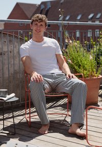 Czerwone metalowe krzesło, na którym siedzi osoba w białej koszuli i w paskowane spodnie. Drewniana podłoga, doniczkowe rośliny i mały czarny stolik w tle.