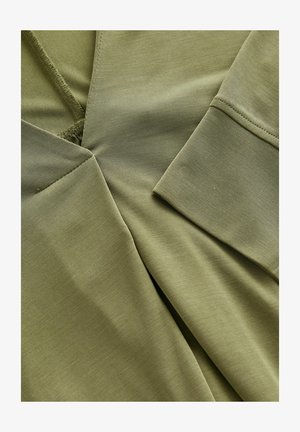 Olivovo zelená látka s hladkou štruktúrou a viacerými vrstvami. Disponuje výstrihom do V a preloženými manžetami.