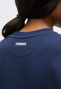 Personne aux cheveux foncés attachés portant une chemise bleu marine avec le mot « STRONGER » imprimé dans le haut du dos.