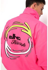 Helle pinke Jacke mit hohem Kragen, die mit schwarzen und neon-gelben grafischen Spiralen verziert ist und den Text "Elho Freestyle" auf dem Rücken trägt.