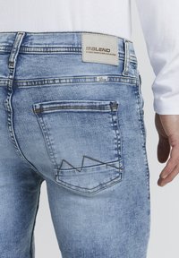 Ljust blå denimjeans med ett distinkt zigzag-sömnadsmönster på bakfickan. Taggen visar varumärket "BLEND."