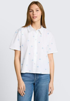 Młoda kobieta w białej koszuli z krótkim rękawem zapinanej na guziki z drobnymi niebieskimi nadrukami muszelek i niebieskich dżinsach, stojąca na jasnym tle.