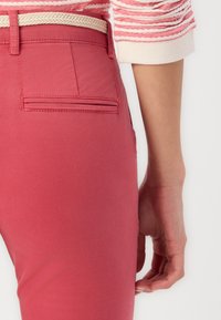 Pantalones de algodón rojos con una textura suave y un bolsillo en la parte trasera. Un cinturón de cuerda trenzada complementa el conjunto, que se acompaña de una blusa casual con un patrón a rayas.