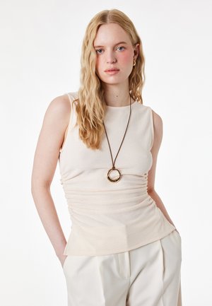 Femme blonde portant un haut beige sans manches, un pantalon crème et un long collier avec un pendentif rond en or, debout les mains dans les poches.