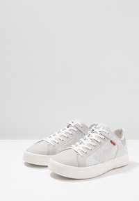 Ljusgrå sneakers med textilöverdel, slät syntetisk tå, vita snören och en röd accentlogotyp på sidan. Flat gummisula.