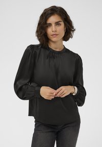 Bluză din satin negru cu guler înalt, mâneci bufante și detalii pliate. Textură netedă și croială relaxată, asociată cu blugi închiși la culoare.