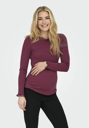 Langsleeve shirt in donker bordeaux met een geribbelde textuur en geaccentueerde manchetten, gecombineerd met zwarte broek, terwijl er met één hand op de heup wordt geposeerd.