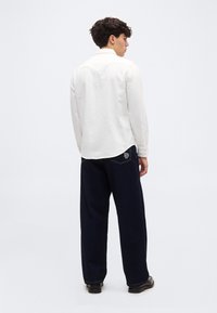 Άνδρας με σκούρα σγουρά μαλλιά, φοράει λευκό πουκάμισο με κουμπιά και φαρδιά σκούρα τζιν με κεντημένη πίσω τσέπη, στέκεται πλαγιαστά.