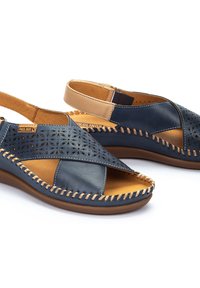 Sandalias de cuero azul marino con diseños perforados, acentos en cuero marrón, bordes cosidos y una correa trasera. Suela cómoda y ligera.