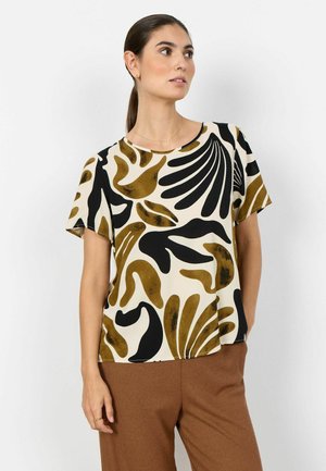 Mujer con cabello oscuro recogido en una coleta, vistiendo una blusa crema con formas abstractas negras y marrones y pantalones marrones, mirando hacia la derecha.