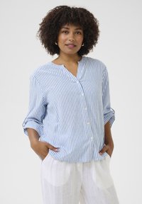 Chemise boutonnée à rayures bleues et blanches avec col mandarin, manches longues retroussées, associée à un pantalon blanc léger.