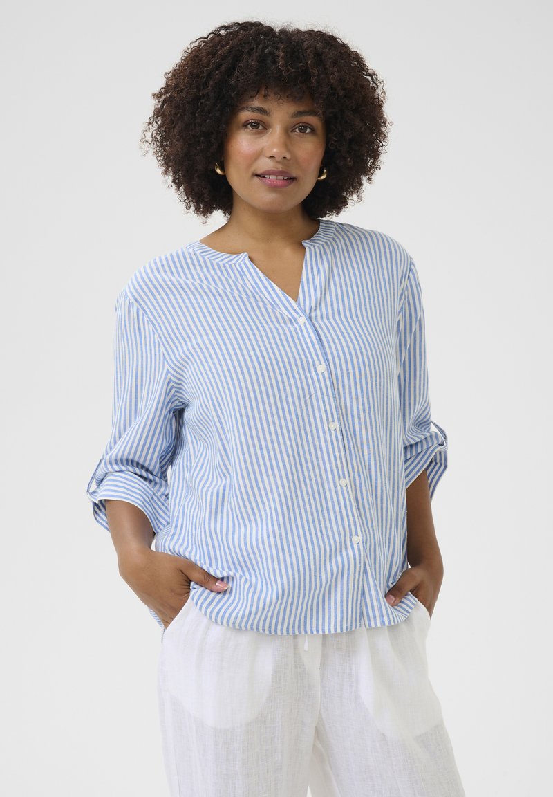 Chemise boutonnée à rayures bleues et blanches avec col mandarin, manches longues retroussées, associée à un pantalon blanc léger.