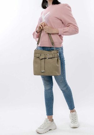 Femme portant un sweat-shirt rose et un jean bleu, tenant un sac à main beige avec un détail de ceinture par les anses.