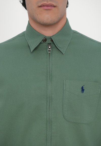 Camisa verde con cremallera y cuello, que cuenta con un bolsillo delantero y un pequeño logo bordado en azul en el pecho. Tejido suave con un acabado mate.