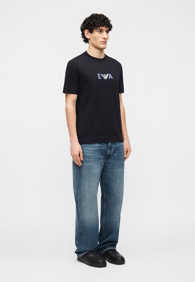 Νέος άνδρας που στέκεται, φοράει μαύρο t-shirt με το λογότυπο EVA, φαρδιά μπλε τζιν και μαύρα αθλητικά παπούτσια, μπροστά σε απλό φωτεινό φόντο.