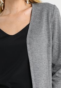 Cardigan grigio con scollatura a V, realizzato in tessuto morbido. Cuciture evidenti e uno stile pulito e minimalista. Indossato sopra una maglietta nera.