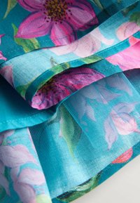 Artimųjų planas sluoksniuoto žalsvo audinio su rožiniais ir violetiniais gėlių raštais bei permatomo tinklelio detalėmis.