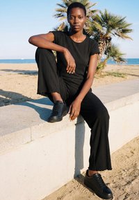 Joven mujer con conjunto negro de canalé y zapatillas sentada en un borde de hormigón en una playa con palmeras y el océano de fondo.