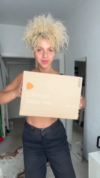 Rjava kartonska škatla z belim napisom "Ljubim me. Nosim me." in logotipom Zalando, ki jo drži oseba v črnih hlačah proti svetli steni.