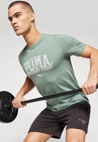 Grünes Baumwoll-T-Shirt mit einem weißen "PUMA Sports Club"-Grafik, kombiniert mit schwarzen Shorts, die ein weißes "HYROX"-Logo tragen. Modell hebt eine Langhantel.