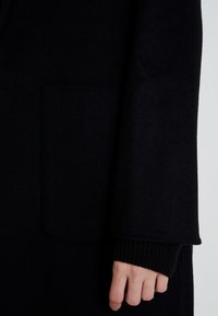 Černý vlněný kabát s hladkou strukturou, s našitou kapsou a žebrovaným lemem na konci rukávu. Jednoduchý design, bez viditelných vzorů.