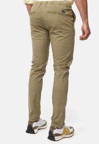 Chinos kaki avec une coupe slim, dotés de deux poches arrière avec zippers et d'une taille élastique. Associés à des baskets blanches et grises.