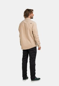 Helle beige Langarmhemd mit Kragen, Knopfmanschetten und einem kleinen Logoetikett auf der Rückseite, kombiniert mit schwarzen Hosen und grünen Sneakers.
