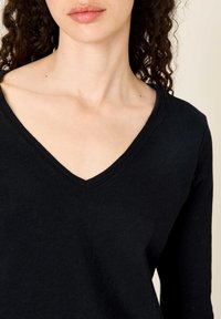 Ženska s kodrastimi temnimi lasmi, oblečena v črno dolgoročno majico z V-izrezom, prikazana od ramen do ustnic na bež ozadju.