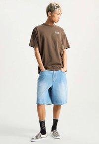 Brązowa, oversize'owa koszulka z logo, jasnoniebieskie szorty jeansowe, czarno-białe slipony w kratę oraz czarne skarpetki z logo.