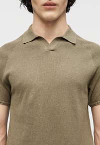 Κοντινό πλάνο ενός ατόμου που φοράει πλεκτή πόλο μπλούζα με κοντά μανίκια σε ελιά απόχρωση, με χωριστό λαιμόκοψη και γιακά.