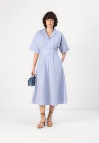 Abito midi a righe blu e bianche con maniche corte, vita cintrata, chiusura a bottoni e tasche. Indossato con scarpe con tacco beige con cinturini e una borsa blu.