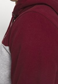 Sudadera de mezcla de algodón en burdeos y gris con un bolsillo canguro y capucha ajustable con cordón. Textura suave con un diseño en bloques de color.