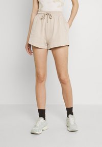 Shorts beiges extensibles avec une ceinture élastique et un cordon de serrage. Comprennent des poches latérales inclinées et une coupe décontractée. Portés avec des baskets vert menthe.