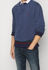 Man som bär en marinblå sweatshirt med röd logotyp över mörka jeans, med ena handen i fickan, stående mot en enfärgad bakgrund.