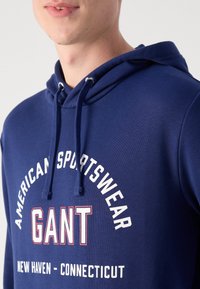 Sudadera con capucha azul marino con un logo en el frente que dice "GANT" en letras blancas. Presenta una capucha con cordones y una tela suave y texturizada.