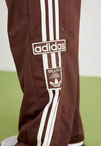 Hnedé teplákové nohavice Adidas s bielymi trojými pruhmi a značkovými nášivkami po stranách, s viditeľnými príchytkami.