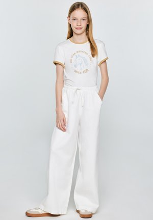 Młoda dziewczyna stoi ubrana w białe spodnie z szerokimi nogawkami, biały t-shirt z beżową lamówką i nadrukiem konia oraz beżowo-białe trampki.