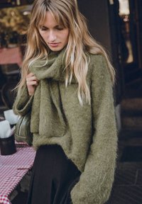 Oliwkowy, puszysty sweter o luźnym kroju i szerokich rękawach, w zestawieniu z ciemną spódnicą. Na lewym boku widoczna torebka kopertówka.