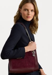 Bordeauxfarbene Lederhandtasche mit strukturiertem Finish, silberner Kettenriemen und einem R.L.-Logo-Anhänger. Ihre Form ist rechteckig mit abgerundeten Ecken.